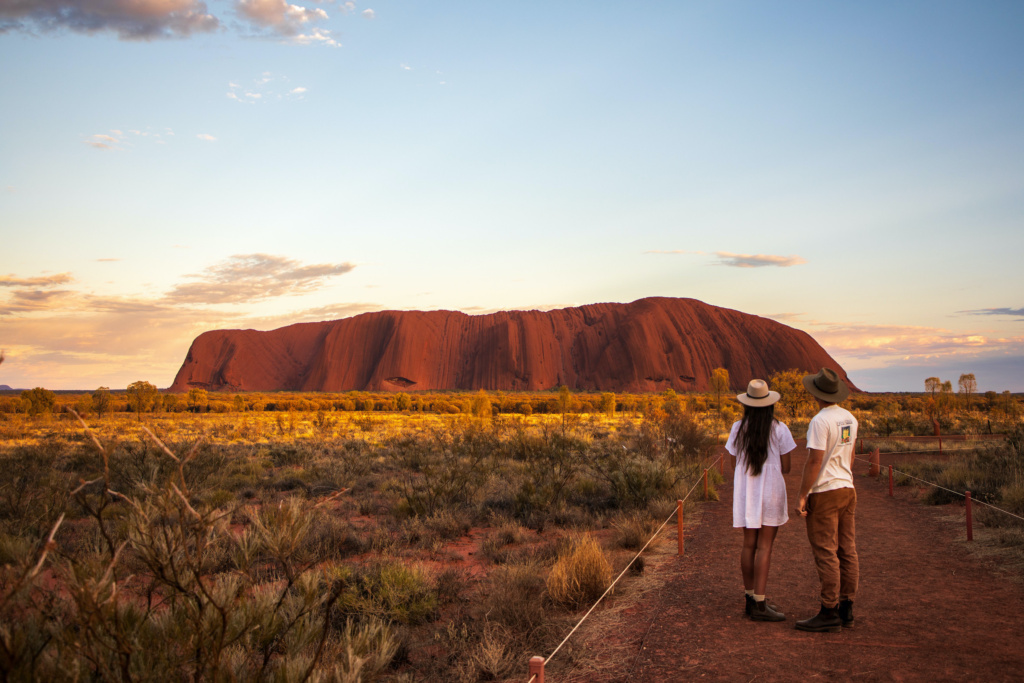 A landscape image of Uluru. Two visitors  are in the foreground of the image on a path that is surrounded by short grass. They are facing away from the camera and looking at Uluru in the distance. 