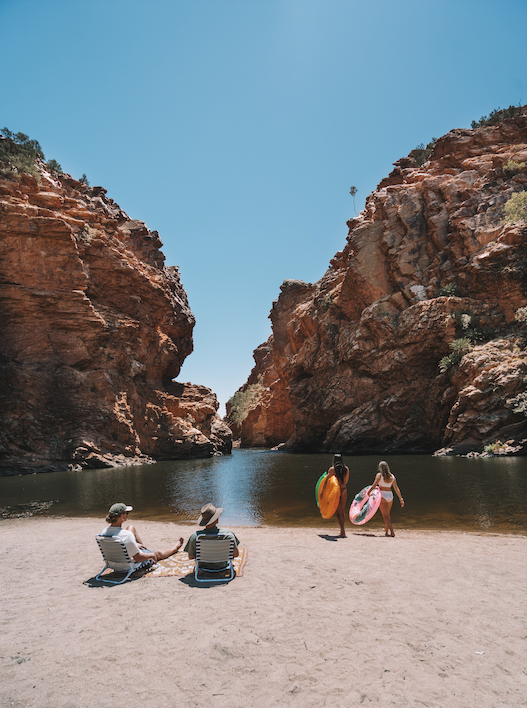 a portrait photo of an opening of water in the West Macdonnell Ranges. There is sand in the foreground of the image with two people relaxing on beach chairs and two others heading into the water. The water has two rocky peaks on either side of it, with an opening in between them revealing the horizon. 