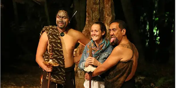 Two Maori men dressed in traditional clothing posing with a smiling tourist who is stood between them.