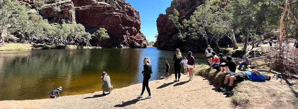 Two Women In Bikinis Lying Down On A Sandy Bank Beside A Body Of Water In Alice Springs