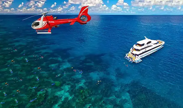 An aerial shot of the Great Barrier Reef. A red helicopter is flying through the air on the left hand side of the shot and a large white cruise boat is anchored in the calm blue waters of the reef on the right. In the lower left of the photo, below the helicopter, a group of snorkellers are exploring the reef.