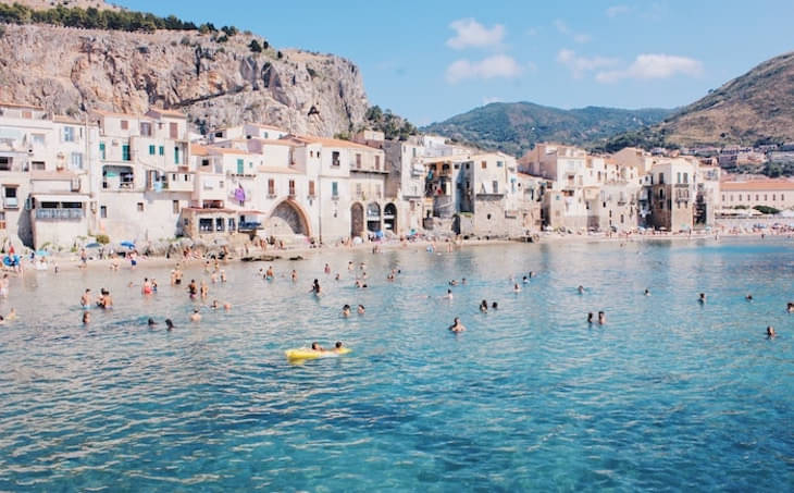People Swimming In Sea In A Sicily, Italy Beach With White Wall And Orange Roof Buildings And Mountain Range Behind