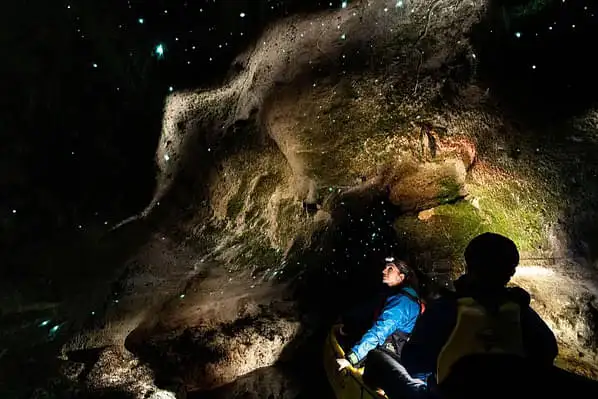 A photo of a person with a headlamp exploring inside a dark cave. The cave's ceiling is speckled with glowing spots caused by the resident glow worms.