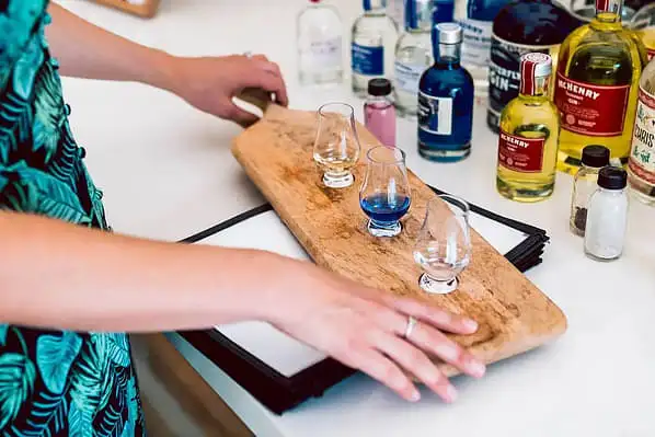 A photograph of a person's hands about to lift a wooden tasting platter of three boutique Tasmanian gins. Many bottles of gin are situated behind the platter.