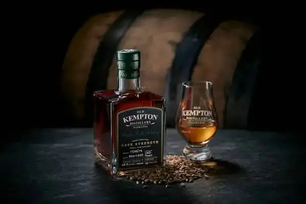 A branded bottle of Old Kempton Distillery whiskey next to a branded glass containing the same whiskey. The bottle and glass are framed in moody lighting on a black table. They are set against a barrel in the background, which is out of focus.